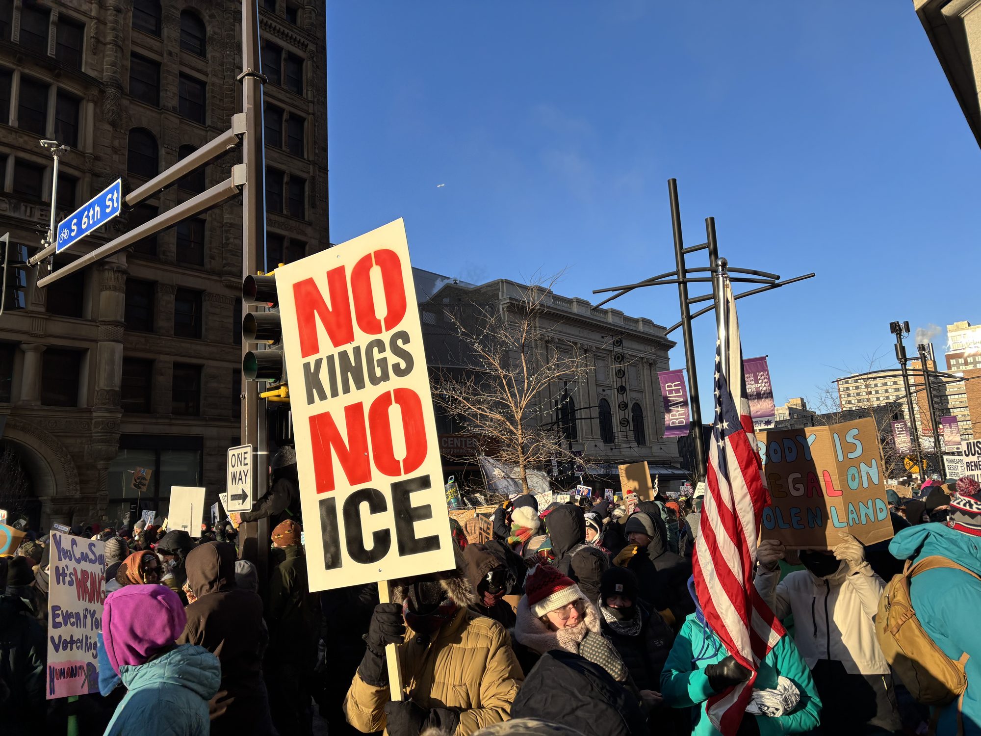 A crowd fills a downtown Minneapolis street on January 23, 2026, with a hand-painted “NO KINGS NO ICE” sign held above the crowd next to an American flag, during protests following Renée Good’s killing by an ICE agent. Photo: Myotus, Wikimedia Commons, CC BY 4.0.